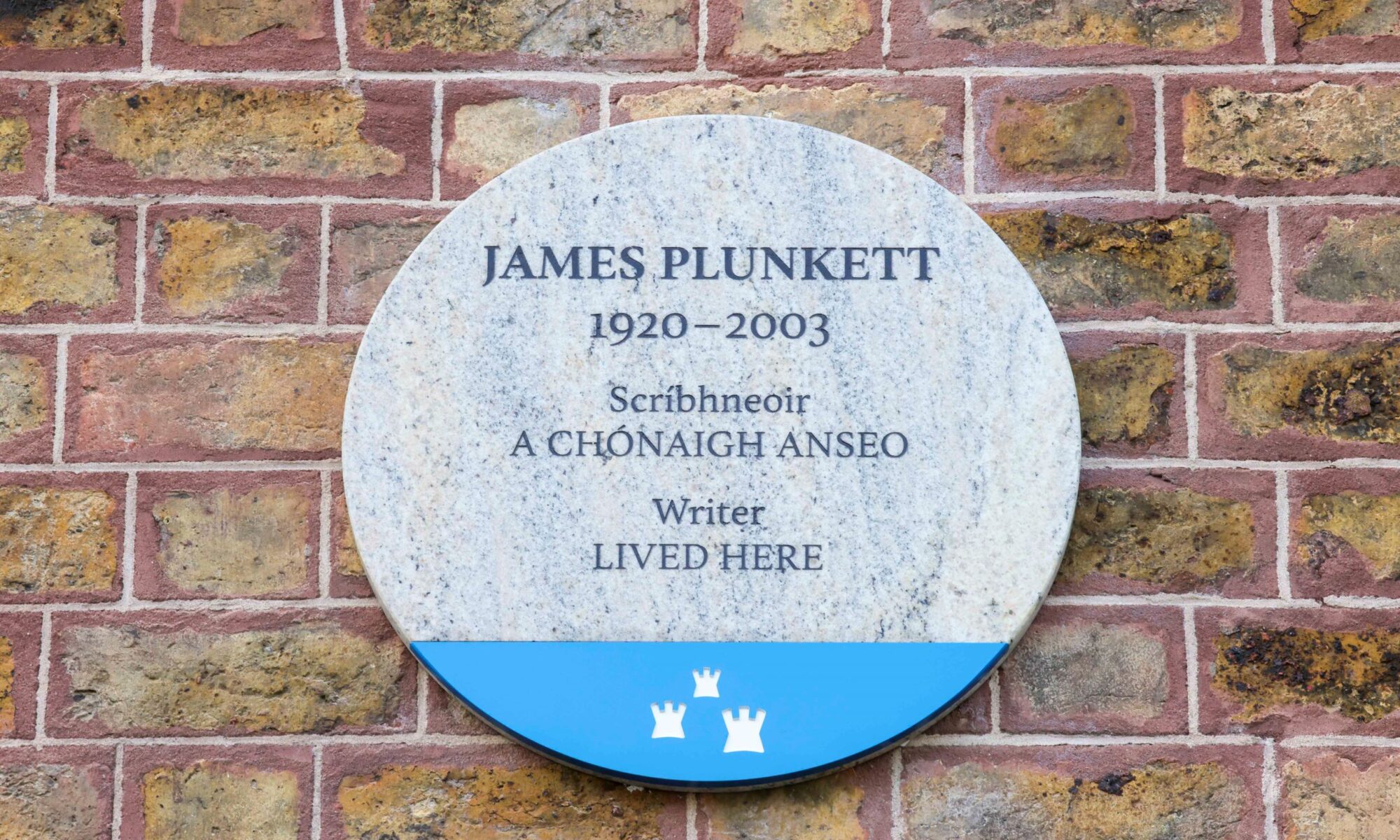 Photograph of a Dublin City plaque. on a red brick wall. The plaque is made of granite and has a blue base with the Dublin City Council logo on it. The text on the plaque is in both Irish and English and in English it reads 'James Plunkett 1920-2003 writer lived here'.
