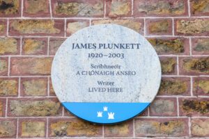 Photograph of a Dublin City plaque. on a red brick wall. The plaque is made of granite and has a blue base with the Dublin City Council logo on it. The text on the plaque is in both Irish and English and in English it reads 'James Plunkett 1920-2003 writer lived here'.
